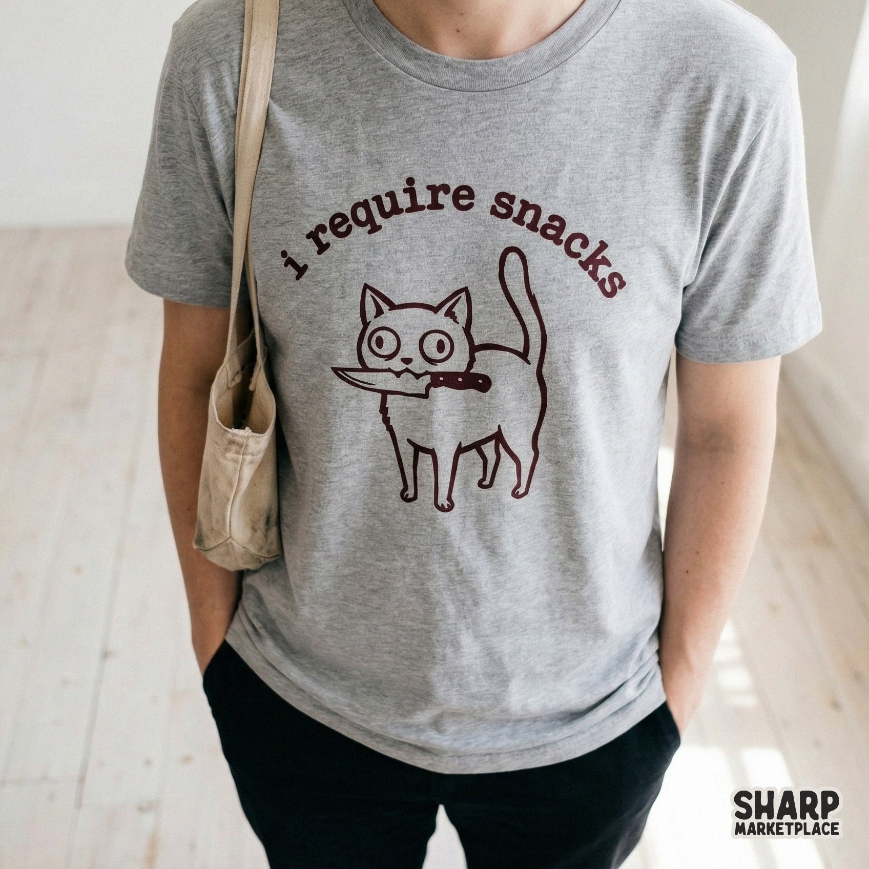 A grey t-shirt featuring a funny illustration of a cat with a knife in its mouth and the text I require snacks in a dark red tone for apparel printing.