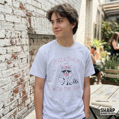 A light gray t-shirt featuring a funny cowboy raccoon illustration with a red bandana and the quote Rizz Them With The Tism for apparel printing.