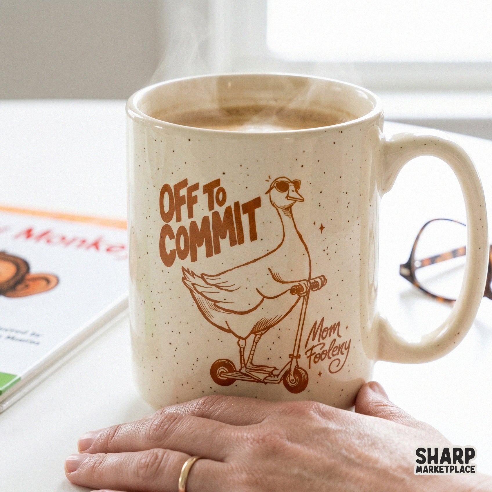 A speckled ceramic coffee mug featuring a brown graphic of a goose on a scooter with the text Off To Commit Mom Foolery for trendy apparel.