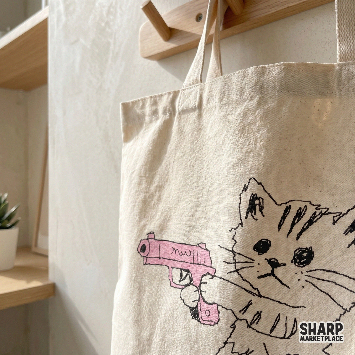 Close up of a whimsical hand drawn kitten illustration with a pink toy gun printed on a canvas tote bag for trendy apparel and DIY craft ideas.