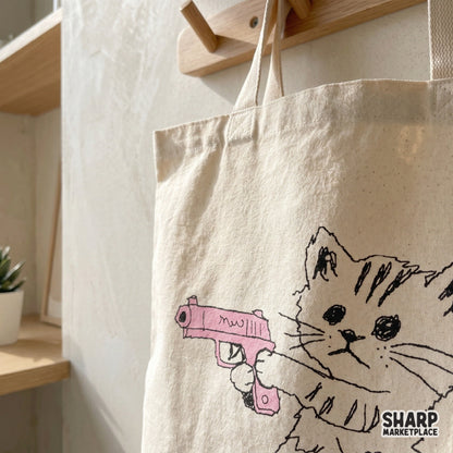 Close up of a whimsical hand drawn kitten illustration with a pink toy gun printed on a canvas tote bag for trendy apparel and DIY craft ideas.