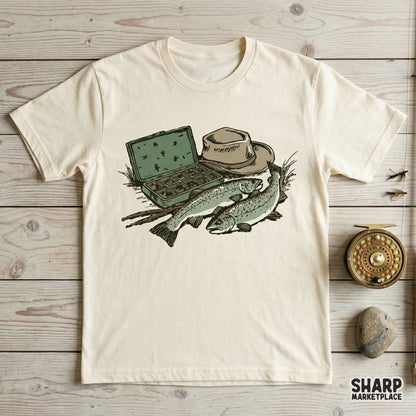 Digital design of trout fishing gear printed on a cream t-shirt, showcasing a fly box, angler hat, and two fish for a rustic outdoor apparel look.