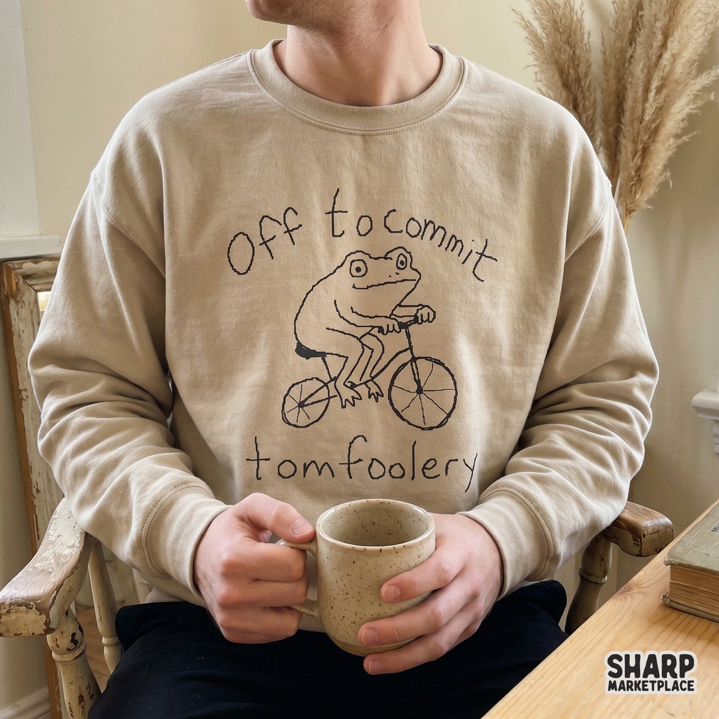 A beige crewneck sweatshirt featuring a black line art illustration of a frog on a bicycle with the quote Off to commit tomfoolery.