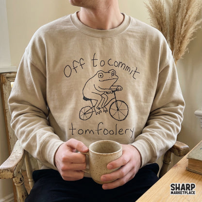 A beige crewneck sweatshirt featuring a black line art illustration of a frog on a bicycle with the quote Off to commit tomfoolery.