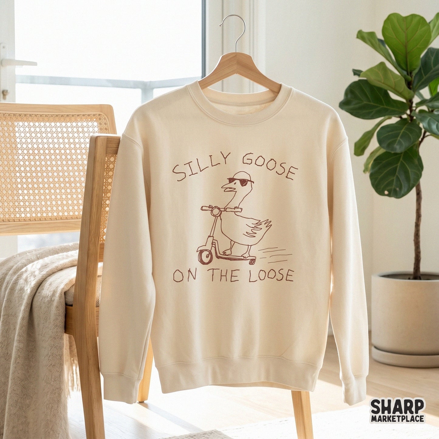 A cream-colored sweatshirt featuring a brown line art illustration of a goose in sunglasses riding a scooter with the text Silly Goose On The Loose.