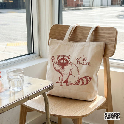 A tote bag featuring a maroon ink design of a cute waving raccoon and the phrase hello there, ideal for custom woodland-themed accessories.