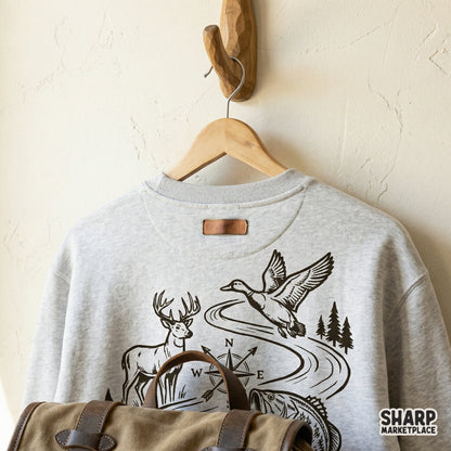 Black ink style outdoor adventure illustration on a gray sweatshirt mockup, featuring a deer, duck, fish, and compass for custom shirt printing.
