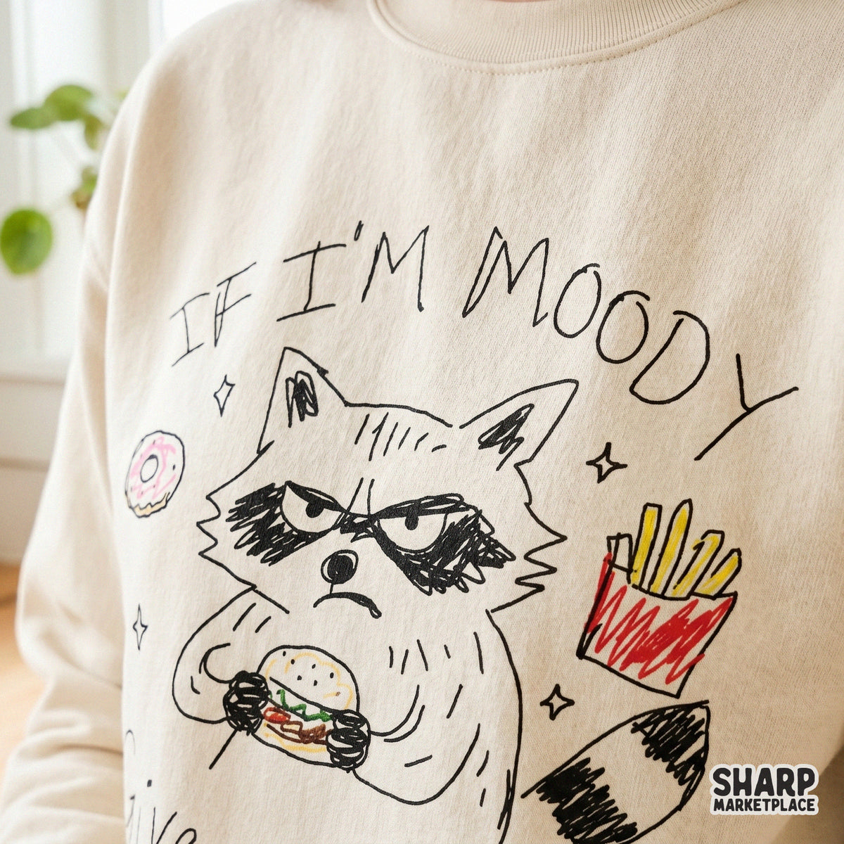 Close-up of a beige sweatshirt featuring a hand-drawn illustration of a scowling raccoon eating a burger with colorful snack accents.