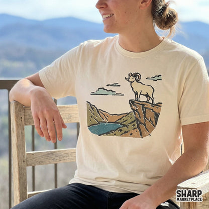 Vintage-style digital graphic of a bighorn sheep on a mountain precipice, displayed on a cream-colored t-shirt for outdoor apparel inspiration.
