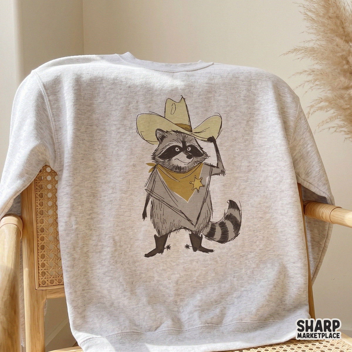 Digital design of a cute raccoon cowboy wearing a tan hat and yellow bandana, featured on a light-colored sweatshirt for country-style clothing