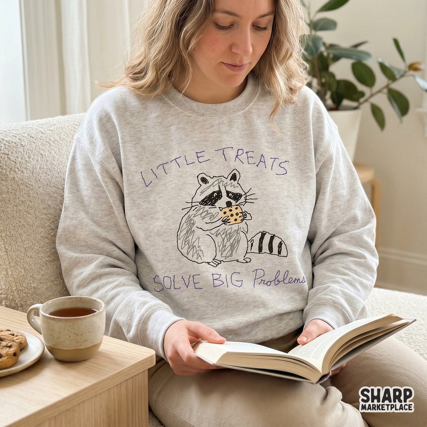 A cozy sweatshirt featuring a whimsical raccoon eating a cookie and the text Little Treats Solve Big Problems, perfect for custom apparel printing.