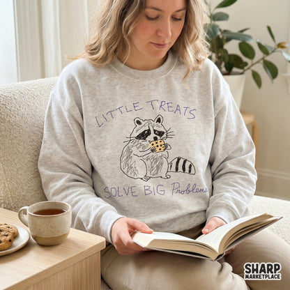 A cozy sweatshirt featuring a whimsical raccoon eating a cookie and the text Little Treats Solve Big Problems, perfect for custom apparel printing.