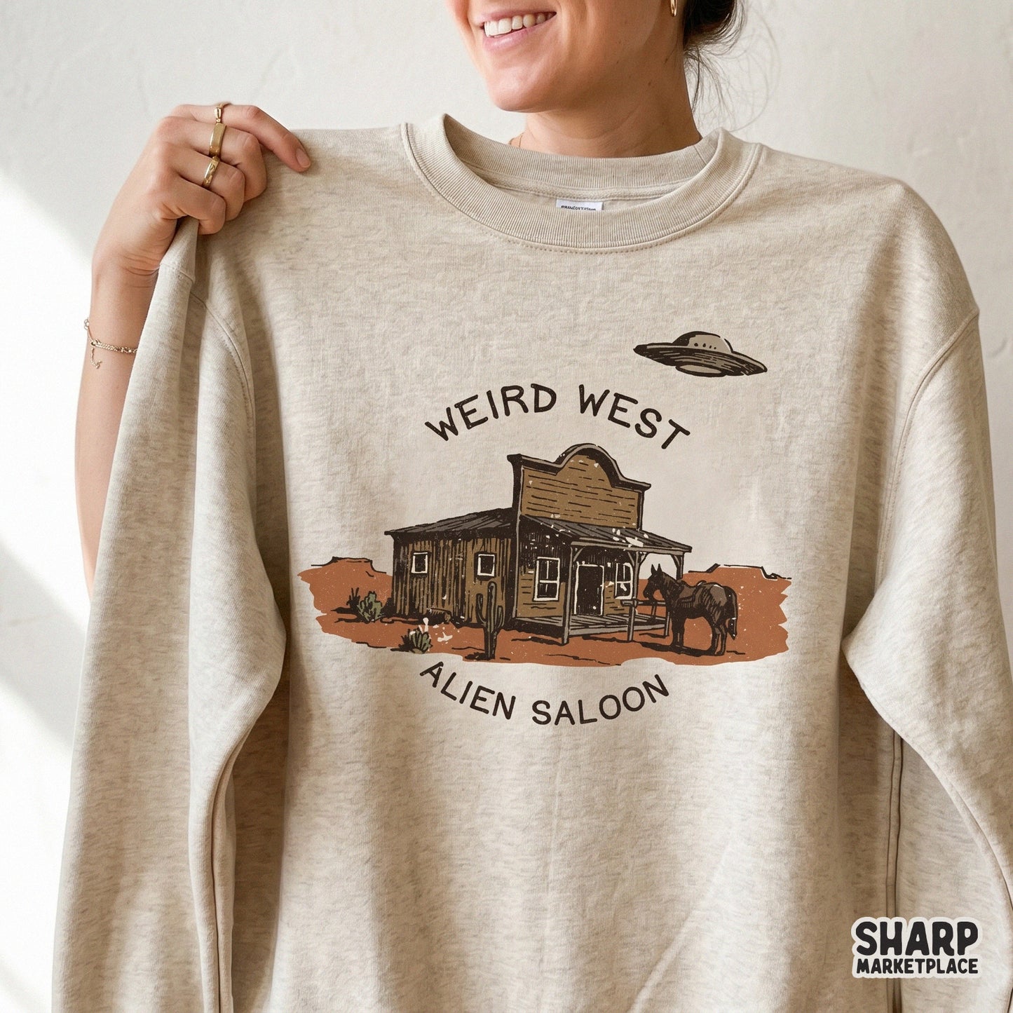A beige sweatshirt featuring a vintage style desert illustration of a wooden saloon, a horse, and a UFO, with the text Weird West Alien Saloon.