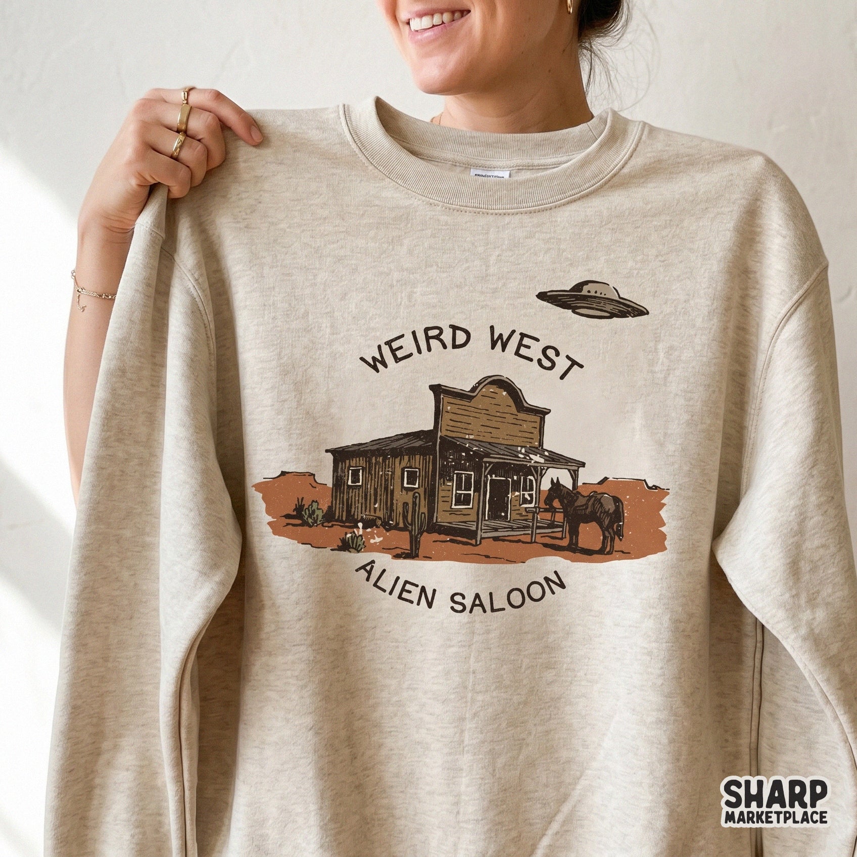 A beige sweatshirt featuring a vintage style desert illustration of a wooden saloon, a horse, and a UFO, with the text Weird West Alien Saloon.
