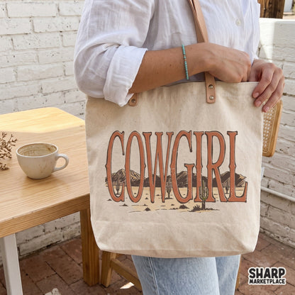 Tote bag featuring a retro western Cowgirl design with desert mountain and cactus landscape graphics inside the bold serif lettering.