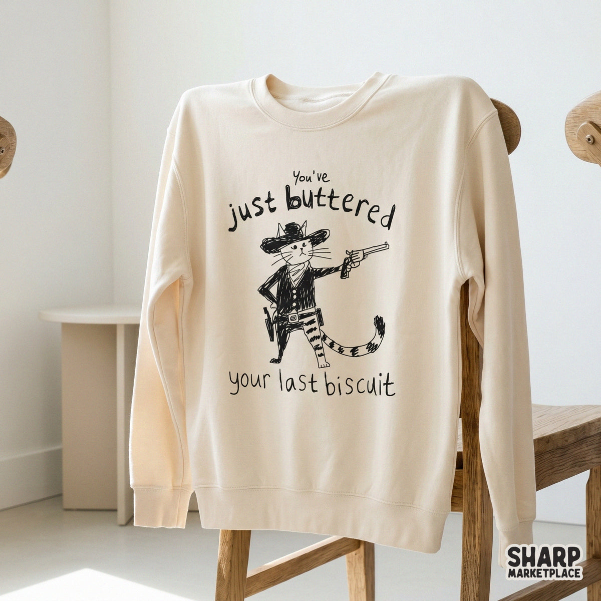 A beige sweatshirt featuring a black ink illustration of an outlaw cat in a western hat and neckerchief, holding a gun with funny sarcastic text.