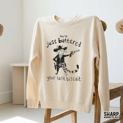 A beige sweatshirt featuring a black ink illustration of an outlaw cat in a western hat and neckerchief, holding a gun with funny sarcastic text.