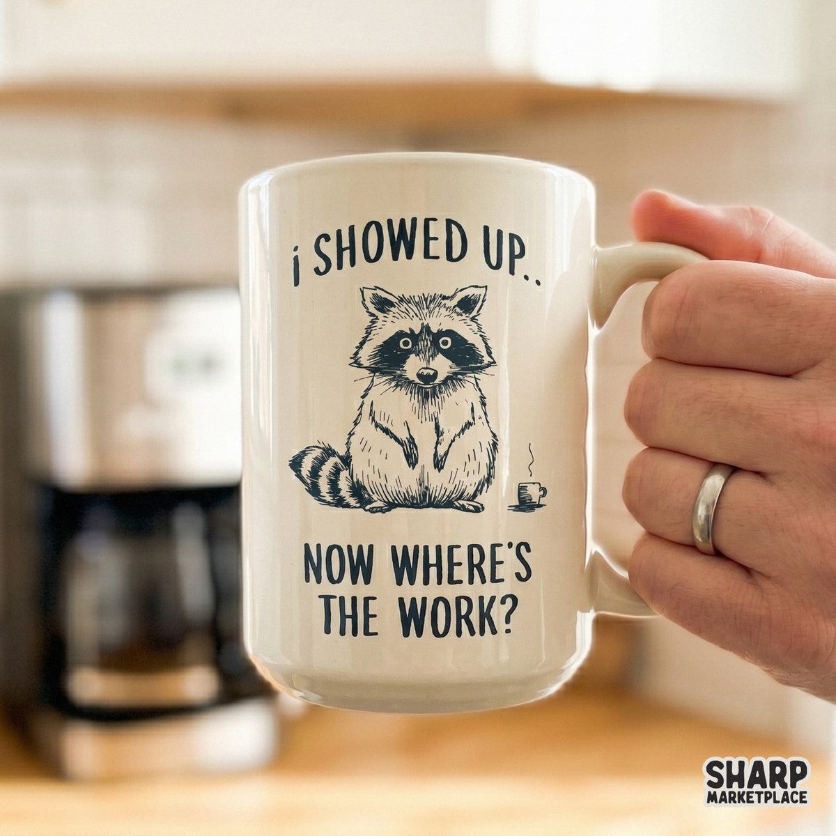 A white ceramic mug featuring a monochromatic illustration of a tired raccoon and the funny office humor quote I Showed Up, Now Where's The Work?