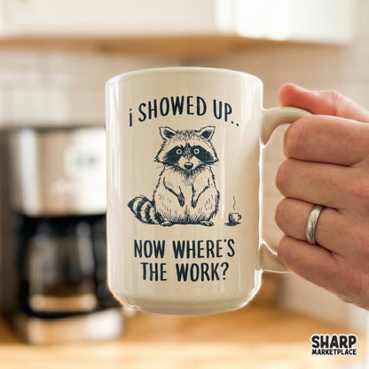 A white ceramic mug featuring a monochromatic illustration of a tired raccoon and the funny office humor quote I Showed Up, Now Where's The Work?