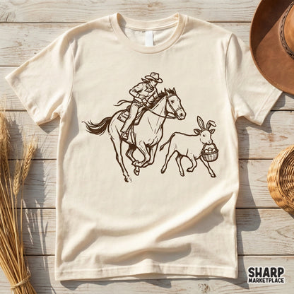 Rustic brown line art design featuring a cowboy riding a horse beside a calf wearing bunny ears and holding an egg basket, printed on a beige t-shirt.