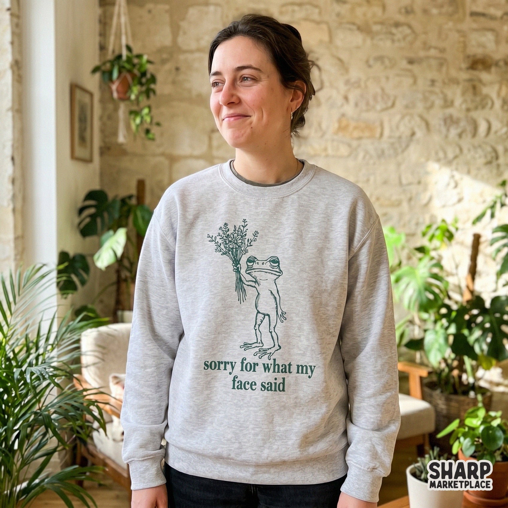 A trendy digital design featuring a disgruntled frog with flowers and the quote sorry for what my face said, displayed on a light grey crewneck sweatshirt.