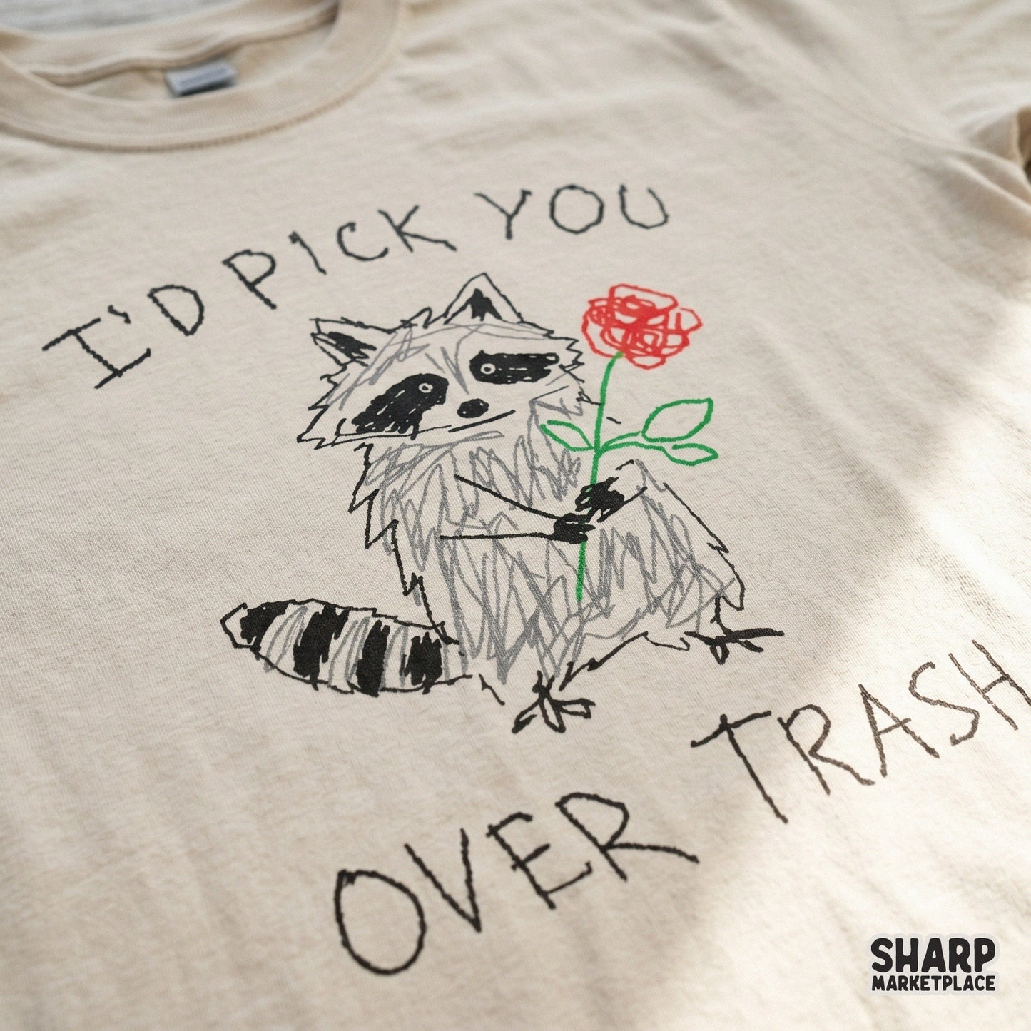 Close-up of a t-shirt design featuring a scribbled raccoon holding a red rose with the funny text I'd Pick You Over Trash for apparel sublimation.