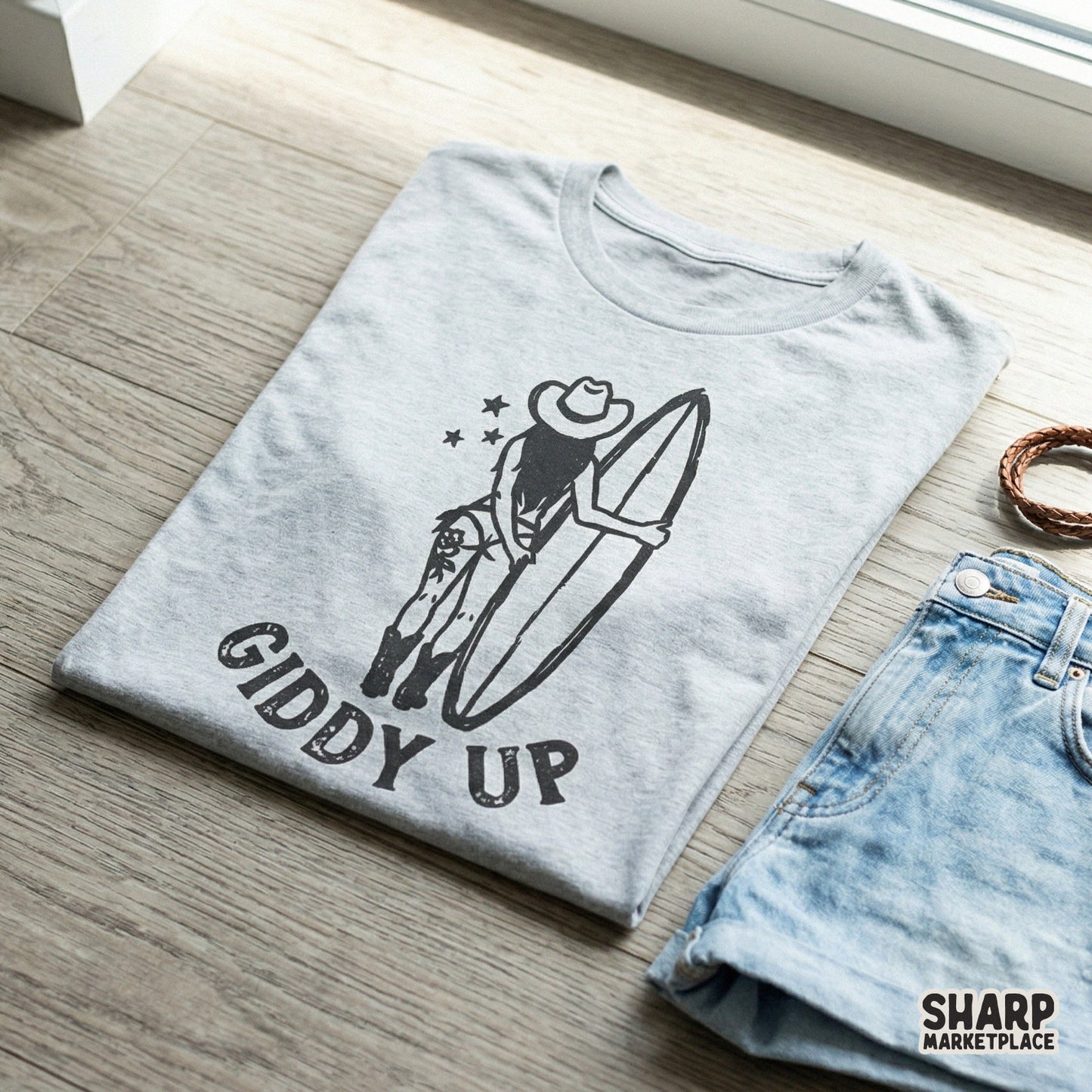 Distressed black ink graphic of a woman in a cowboy hat and boots with a surfboard, shown on a grey t-shirt for coastal rodeo fashion projects.
