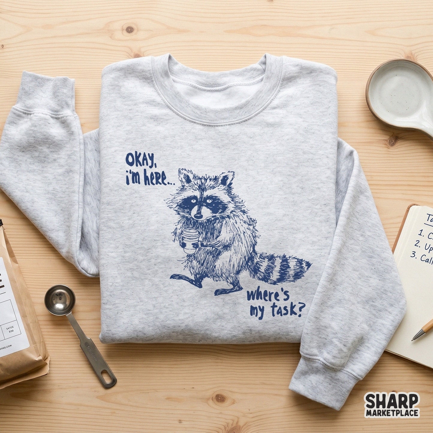 Funny office humor printable graphic featuring a hand-drawn raccoon with a coffee cup and the text Okay, I'm Here... Where's My Task? on a transparent
