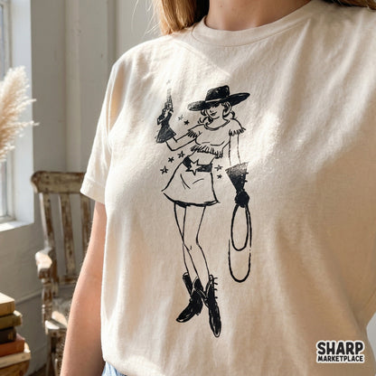 Trendy western cowgirl illustration in a retro pinup style, shown printed on a cream-colored t-shirt for a boutique apparel look.
