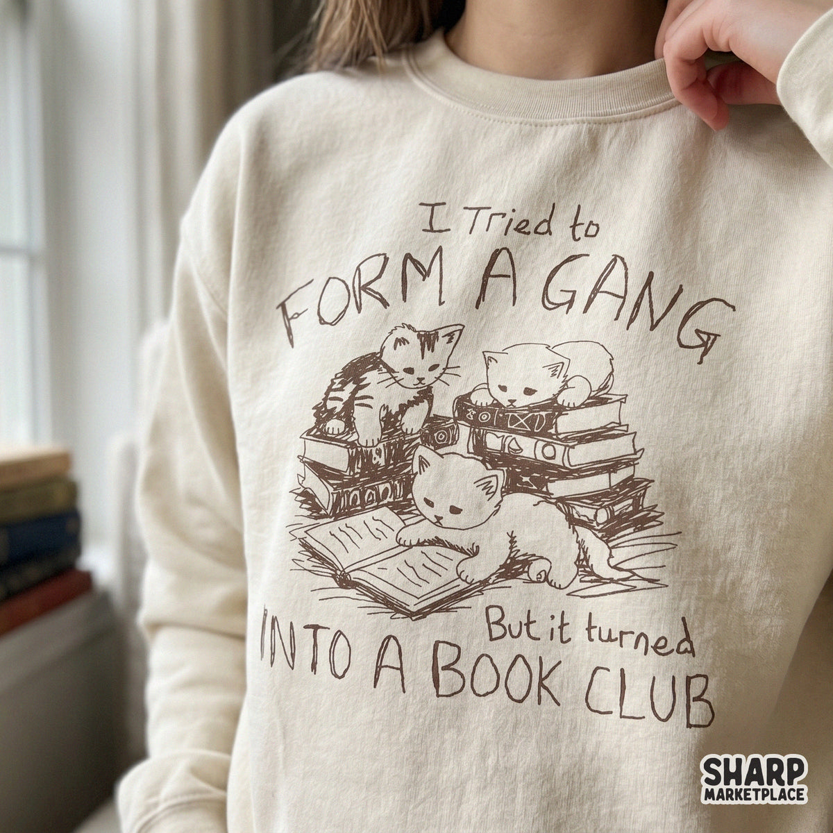 A cozy sweatshirt featuring a brown sketch of three kittens sitting on stacked books with the funny text I Tried To Form A Gang But It Turned Into A Book Club.