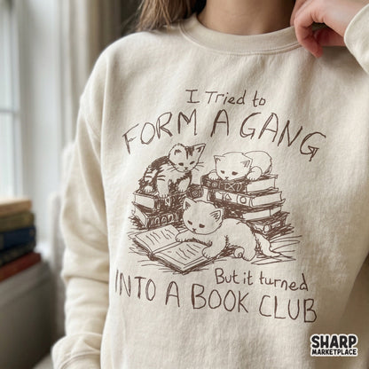 A cozy sweatshirt featuring a brown sketch of three kittens sitting on stacked books with the funny text I Tried To Form A Gang But It Turned Into A Book Club.