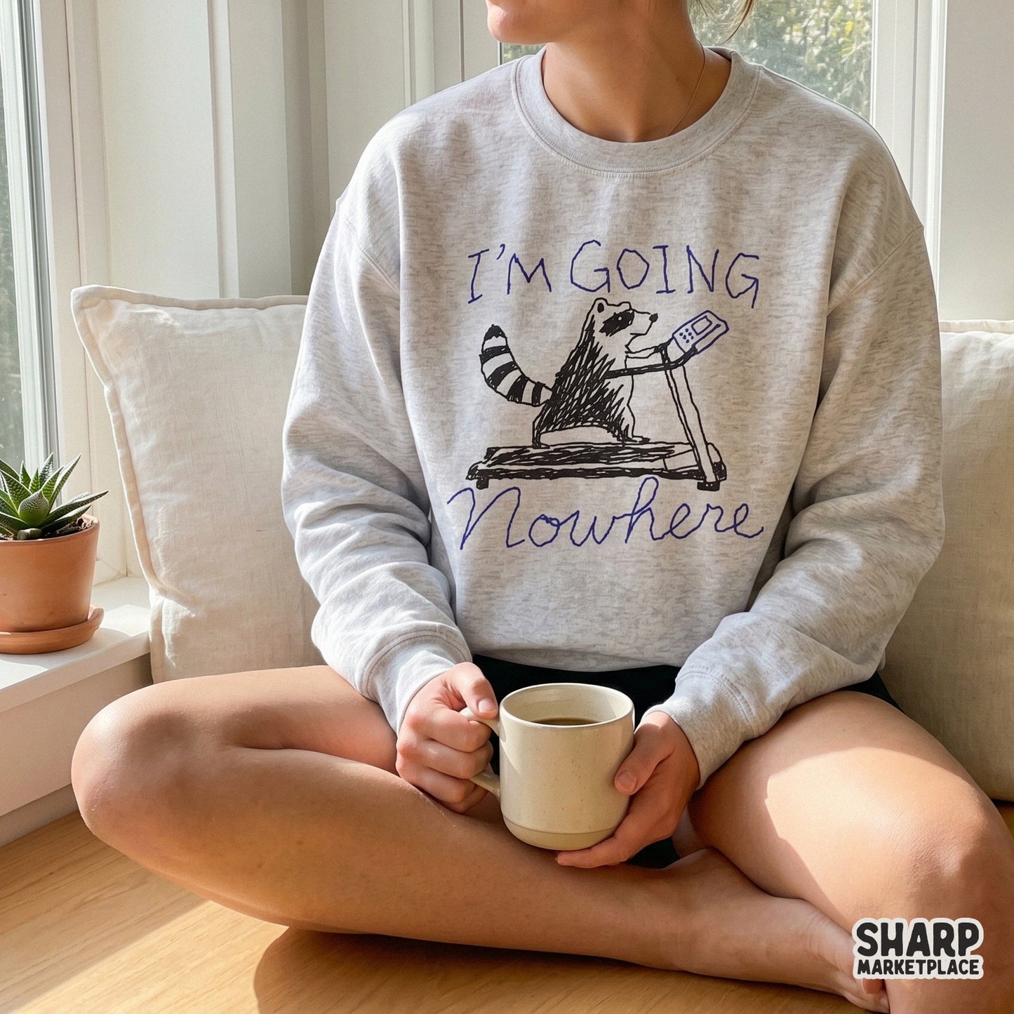 A light gray sweatshirt featuring a hand-drawn illustration of a raccoon on a treadmill with the sarcastic quote I'm Going Nowhere.