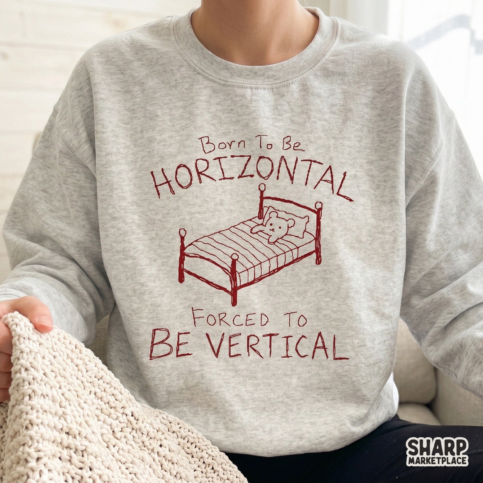 A cozy heather gray sweatshirt featuring a red hand-drawn sketch of a bed with a teddy bear and the sarcastic quote Born To Be Horizontal Forced To Be Vertical.