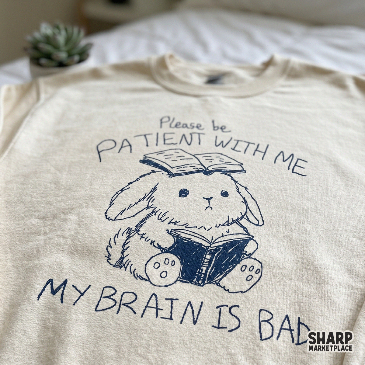 A cream-colored sweatshirt featuring a navy blue doodle of a bunny reading a book with a second book on its head and the sarcastic quote Please be patient with
