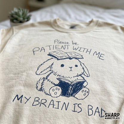 A cream-colored sweatshirt featuring a navy blue doodle of a bunny reading a book with a second book on its head and the sarcastic quote Please be patient with