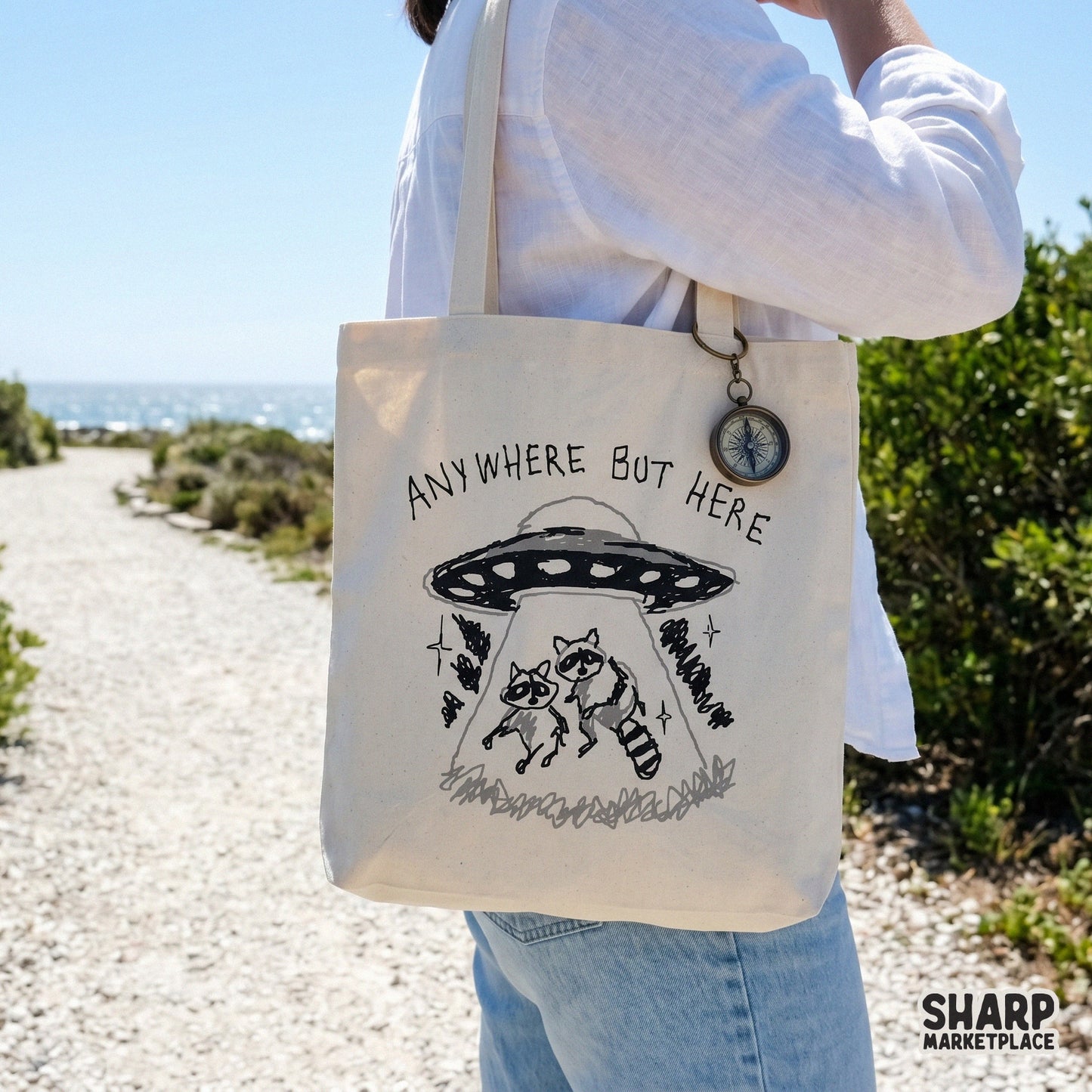 A canvas tote bag featuring a black and white graphic of two raccoons being beamed up by a flying saucer with the sarcastic text Anywhere But Here.