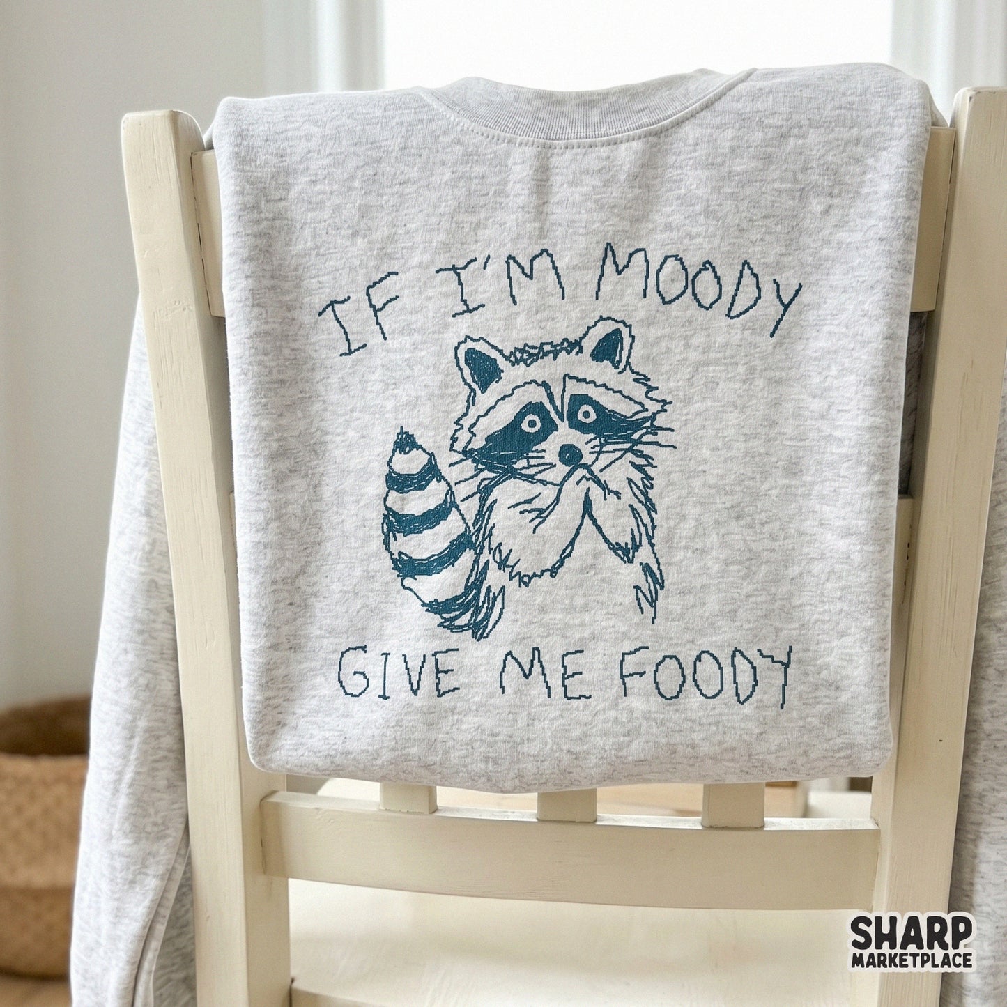 A light grey sweatshirt featuring a dark blue scribbled illustration of a begging raccoon and the humorous quote If I’m Moody Give Me Foody.