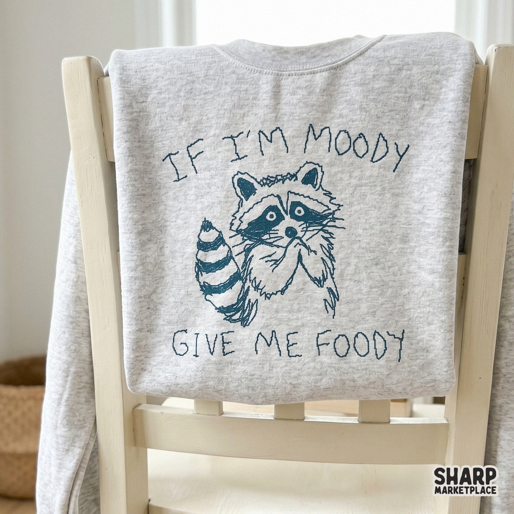 A light grey sweatshirt featuring a dark blue scribbled illustration of a begging raccoon and the humorous quote If I’m Moody Give Me Foody.