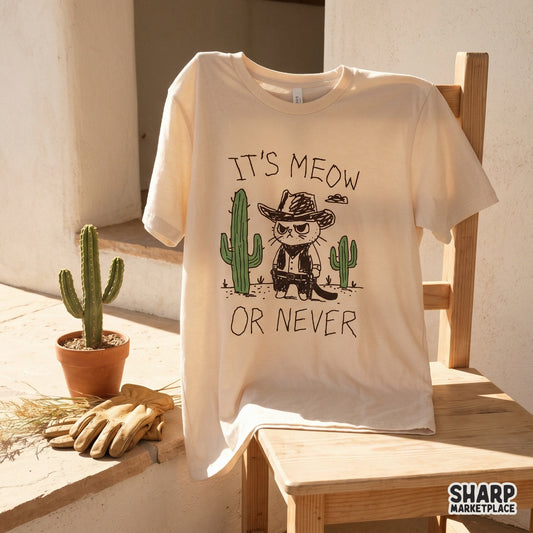 A beige t-shirt mockup featuring a hand-drawn western cat cowboy graphic with green cacti and the funny phrase It's Meow Or Never for apparel printing.