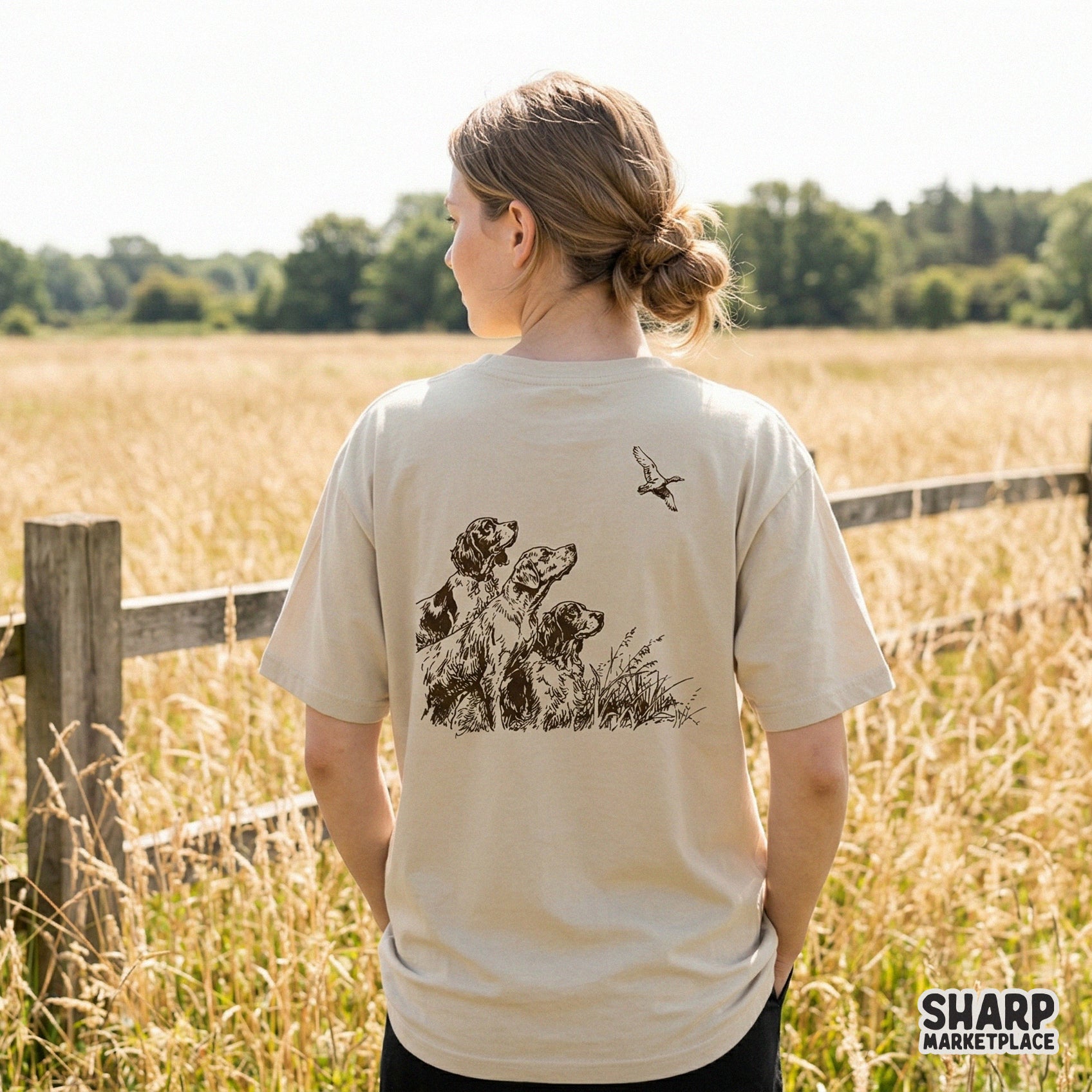 Back view of a beige t-shirt featuring a detailed monochromatic sketch of three hunting dogs watching a duck, ideal for outdoor apparel prints.