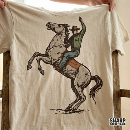 A rustic, hand-drawn illustration of a bronc rider on a bucking horse, displayed on a beige t-shirt mockup for western graphic design inspiration.