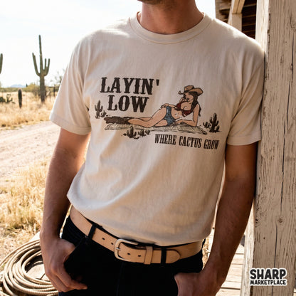 Retro western sublimation design on a beige t-shirt showing a cowgirl relaxing in the desert with cacti, text reads Layin Low Where Cactus Grow.