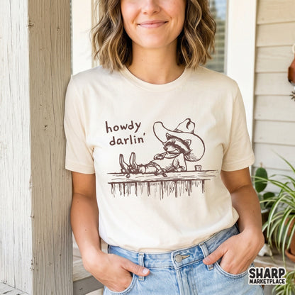 A funny western trash panda wearing a cowboy hat and bandana lounging on a wooden fence with the phrase Howdy Darlin in a retro hand-drawn style.