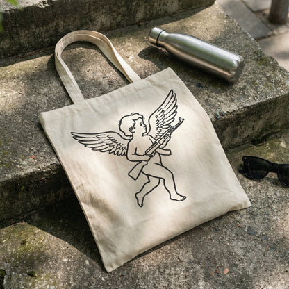 Edgy tattoo-style cherub holding a rifle printed in black ink on a beige canvas tote bag, perfect for streetwear apparel and custom DIY projects.