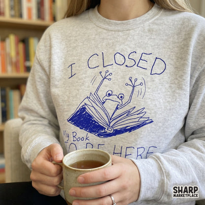 A grey sweatshirt featuring a whimsical blue line art design of a frog emerging from an open book with the quote I Closed My Book To Be Here.