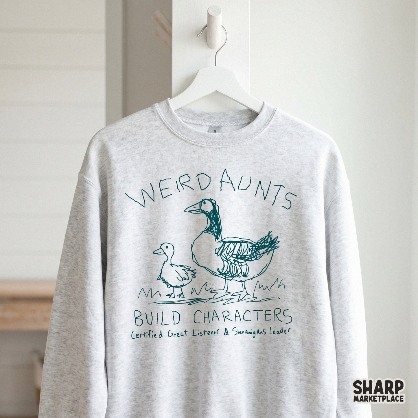 A light gray sweatshirt featuring a teal sketch of a goose and duckling with the humorous quote Weird Aunts Build Characters for apparel printing.