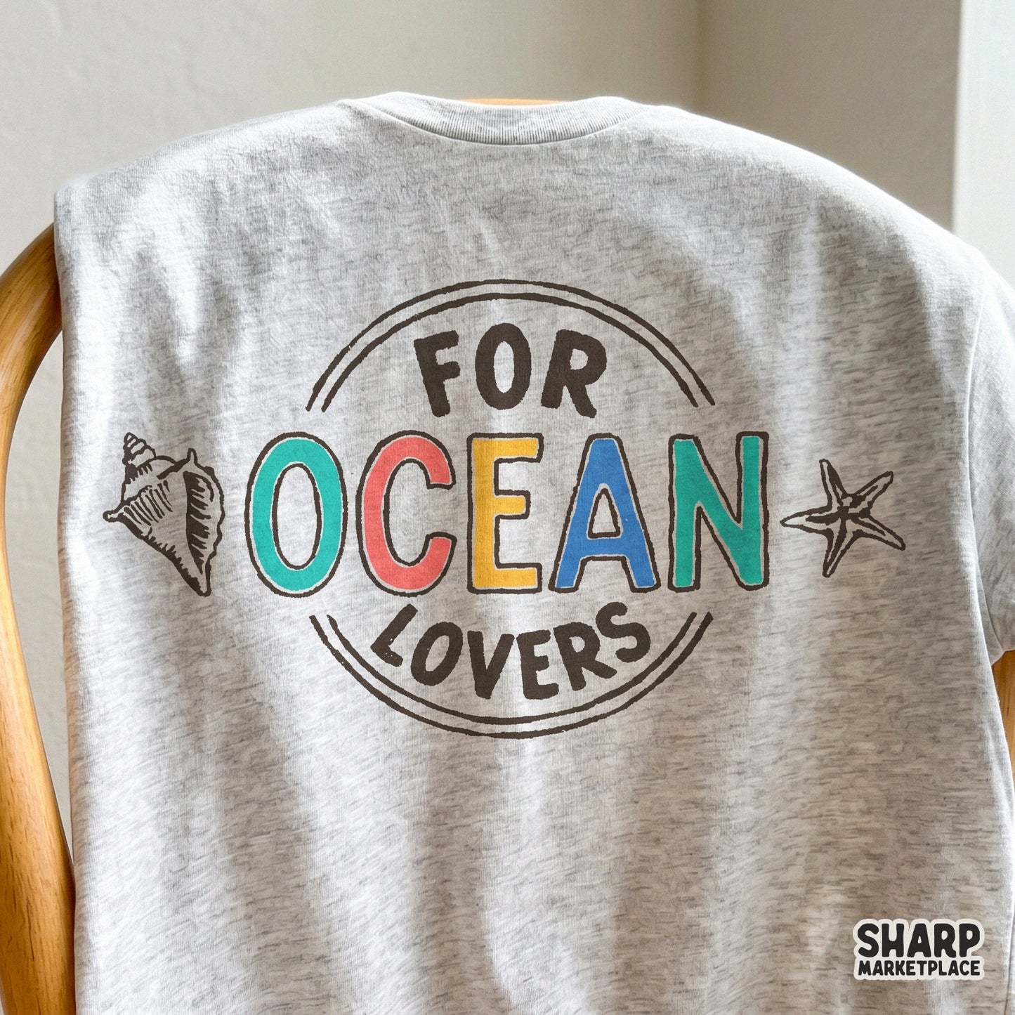 A heather gray t-shirt featuring a colorful For Ocean Lovers coastal design with a circular frame, shell, and starfish for beach-themed apparel.