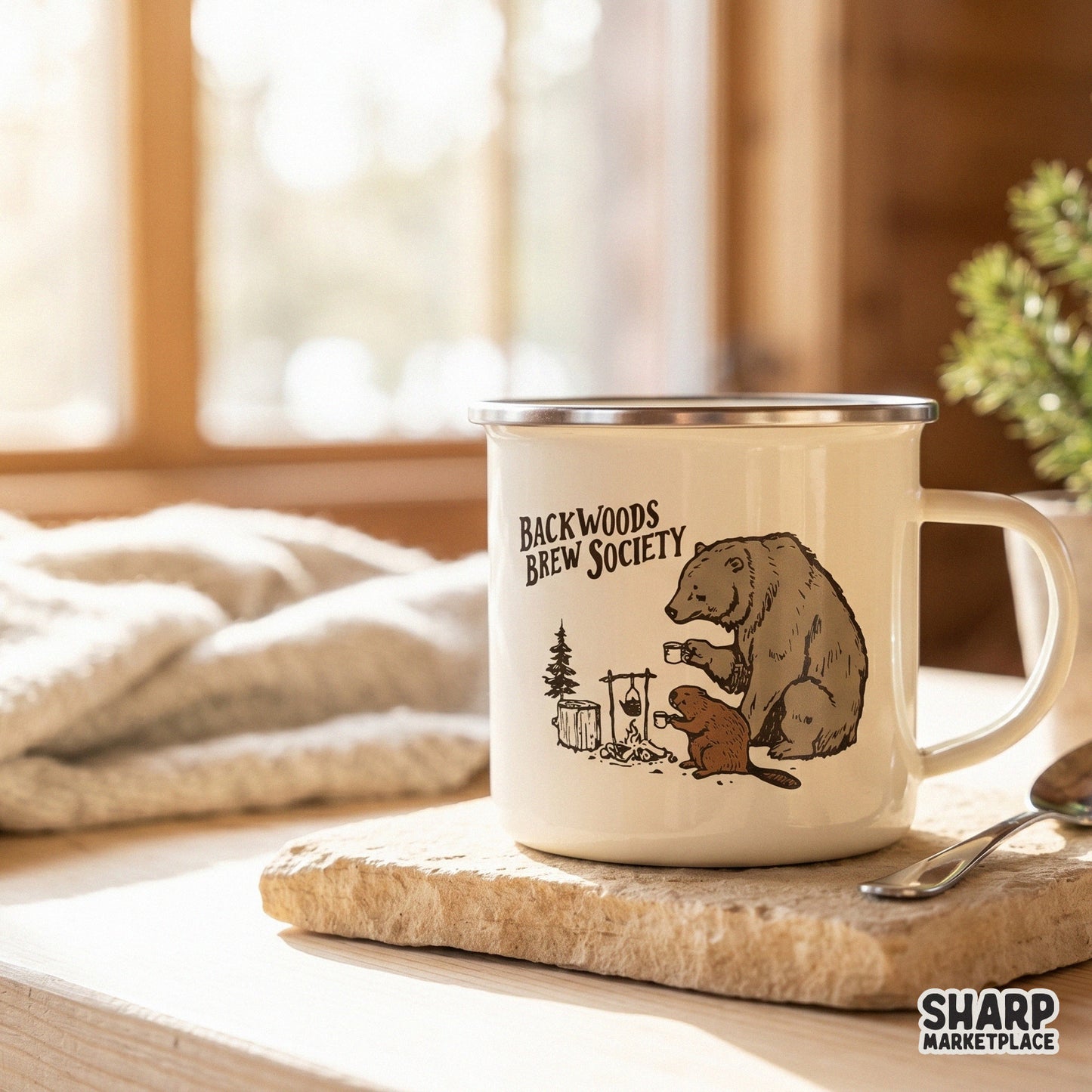 Cream-colored camping mug featuring a rustic illustration of a bear and beaver sharing coffee by a fire with Backwoods Brew Society text.