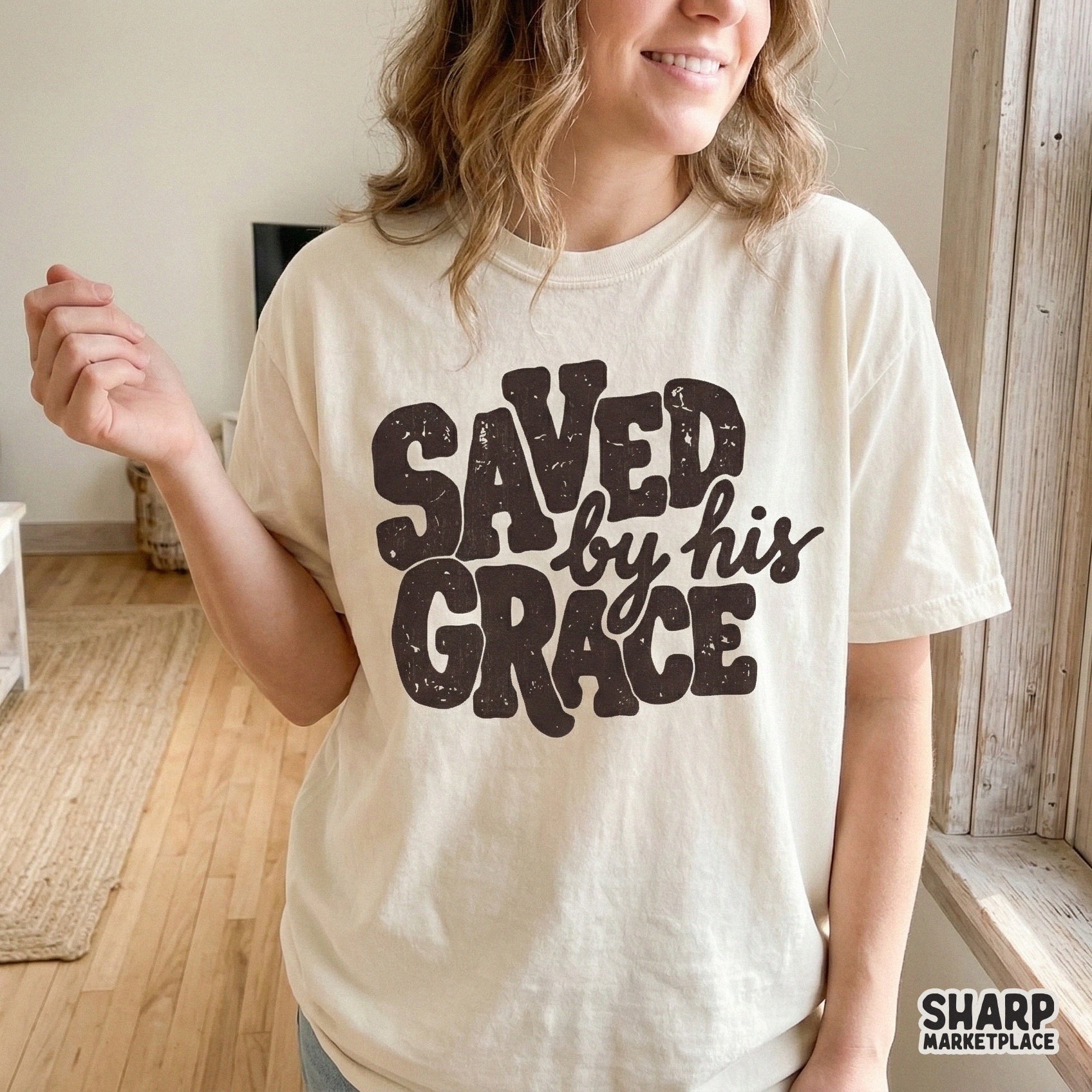 A vintage-style faith graphic featuring bold, textured lettering that says Saved By His Grace, displayed on a neutral t-shirt for apparel inspiration.