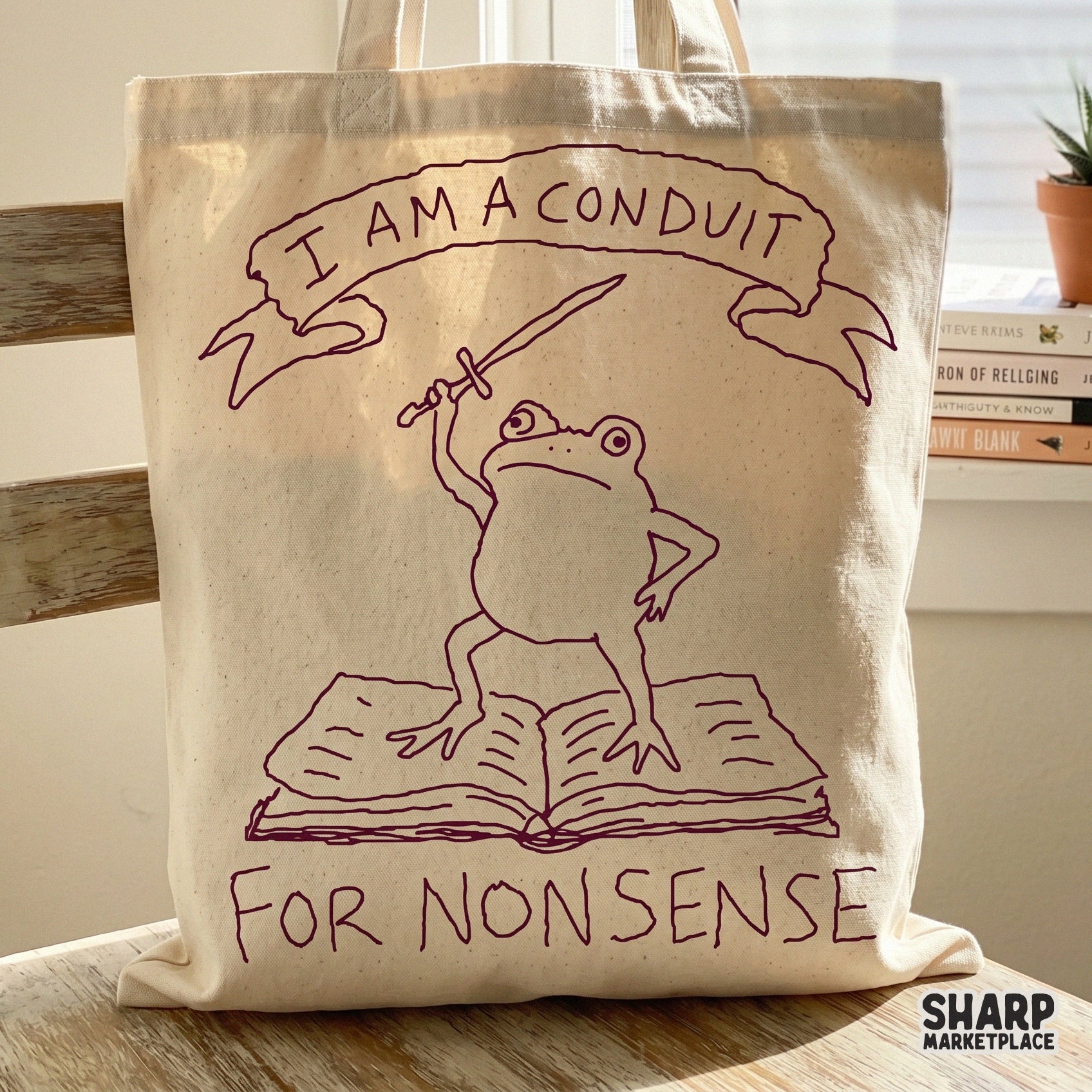 A canvas tote bag featuring a maroon line art design of a frog with a sword standing on a book, captioned I Am A Conduit For Nonsense.
